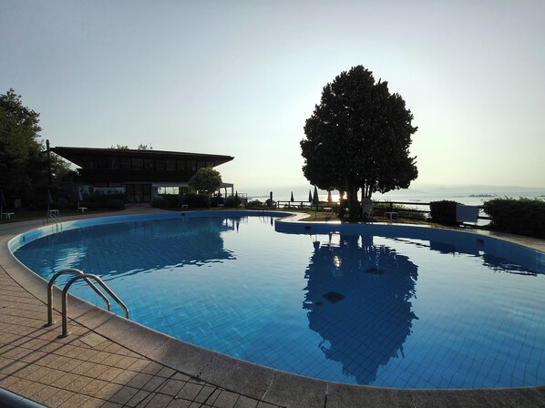 A swimming pool with a clear view of the sea, a large tree, and a building with a sloped roof. - CASA CATULLO, Paradise Children Apartment in Resort (Lombardia)