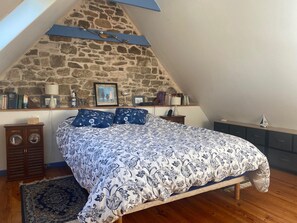 Ein Schlafzimmer mit Steinsandsteinwand, schräg versetzter Decke, einem Bett mit gemusterten Betttextilien, Nachttischen und einem kleinen Teppich.