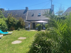 Ein Haus mit Steinaußenfassade, ein kleiner Garten mit Rasenfläche, eine Terrasse mit Gartenmöbeln und ein überdachter Bereich mit Tisch und Stühlen.
