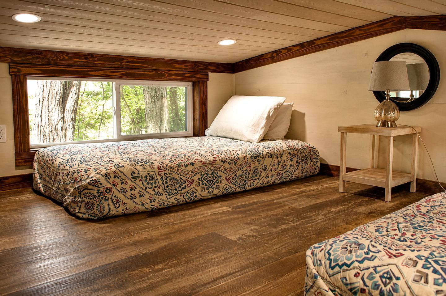 A bedroom with a bed, a nightstand with a lamp, a window with a view of trees, and wooden flooring.