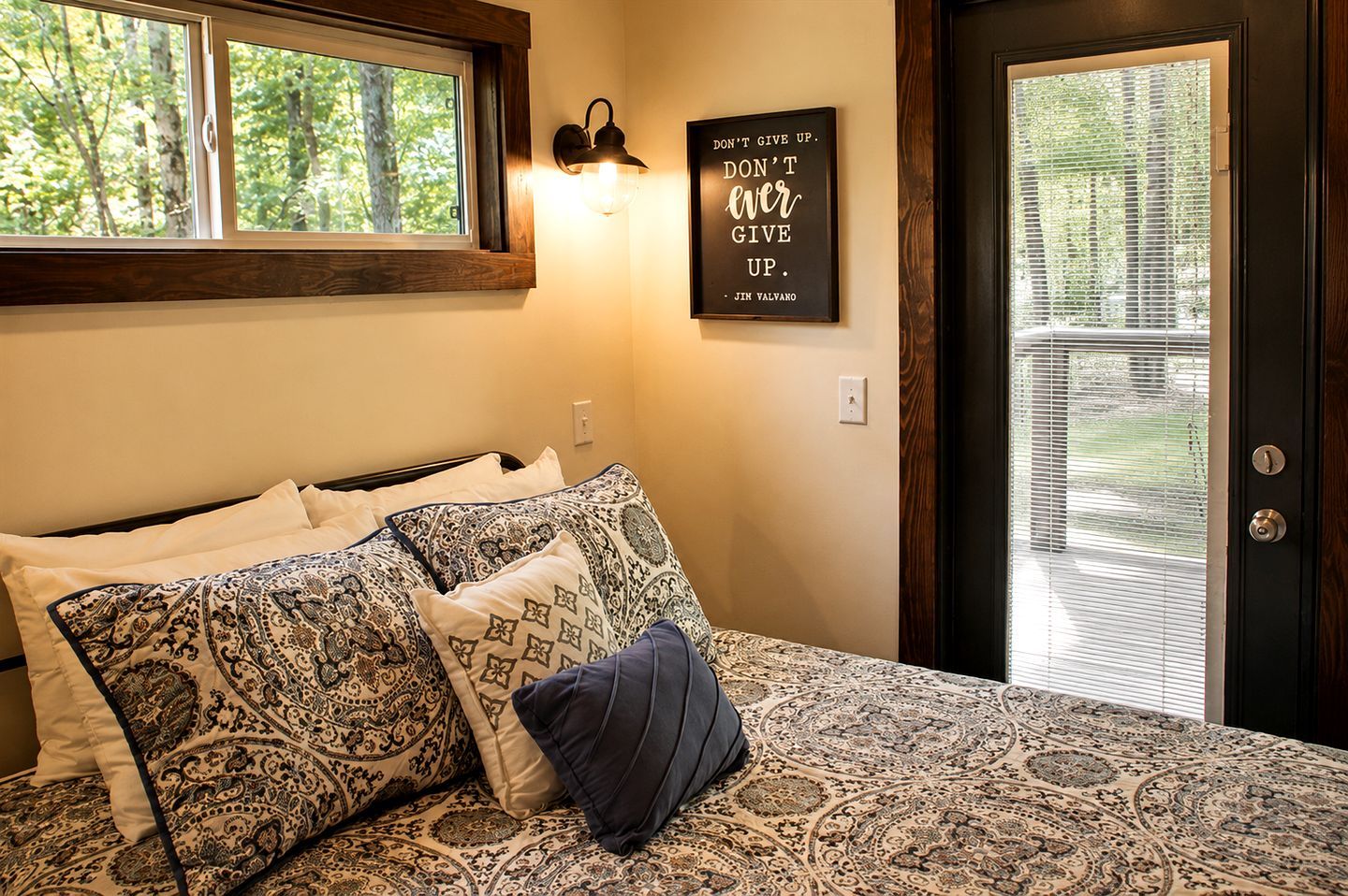 A bedroom with a bed, patterned pillows, a wall-mounted light, and a motivational quote.