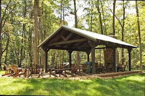 A wooden pavilion with a stone fireplace, surrounded by wooden chairs and tables, set in a forest.