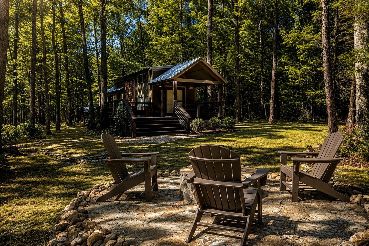 A cabin with a porch, surrounded by a forest. There are two Adirondack chairs in front.