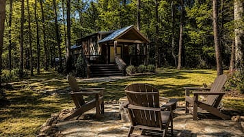 A cabin with a porch, surrounded by a forest. There are two Adirondack chairs in front.