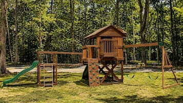 Una estructura de juego de madera con tobogán, muro de escalada y columpios en un parque con césped.
