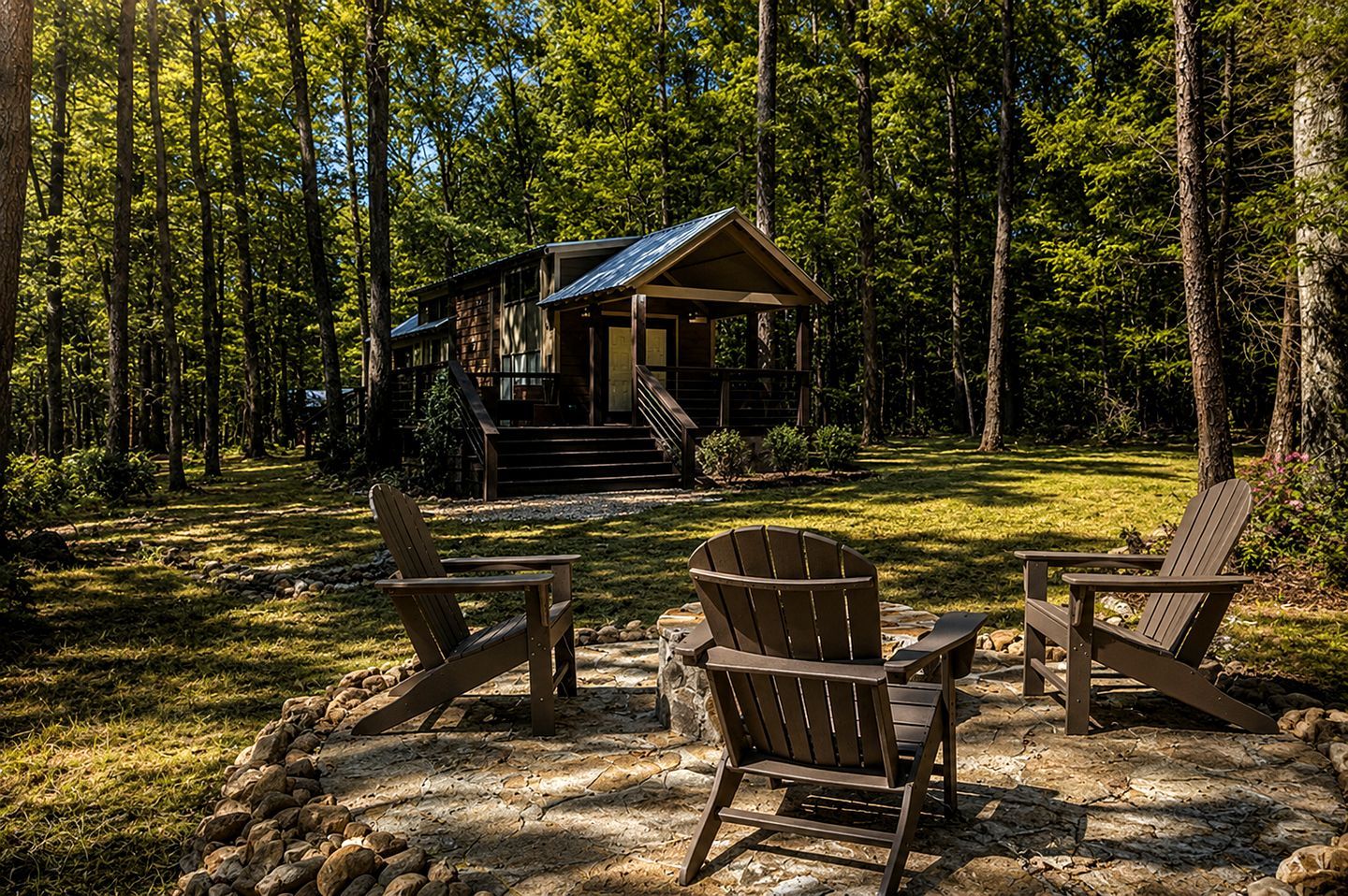 A cabin with a porch, surrounded by a forest. There are two Adirondack chairs in front.