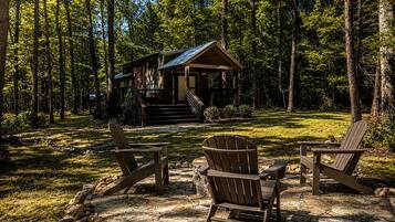 A cabin with a porch, surrounded by a forest. There are two Adirondack chairs in front.