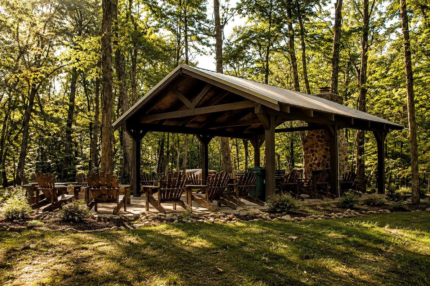 A rustic wooden pavilion with a stone fireplace, surrounded by Adirondack chairs in a forest setting.