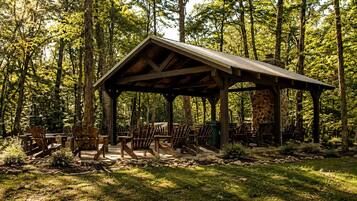 A rustic wooden pavilion with a stone fireplace, surrounded by Adirondack chairs in a forest setting.