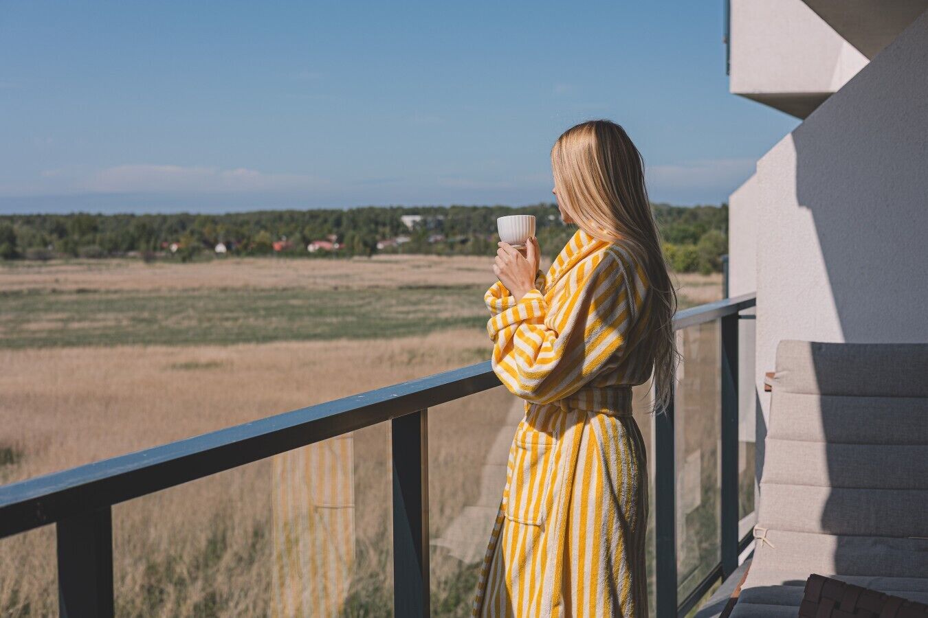 Una persona vestida con una bata a rayas, de pie en un balcón con vistas a un campo y casas al lejano.