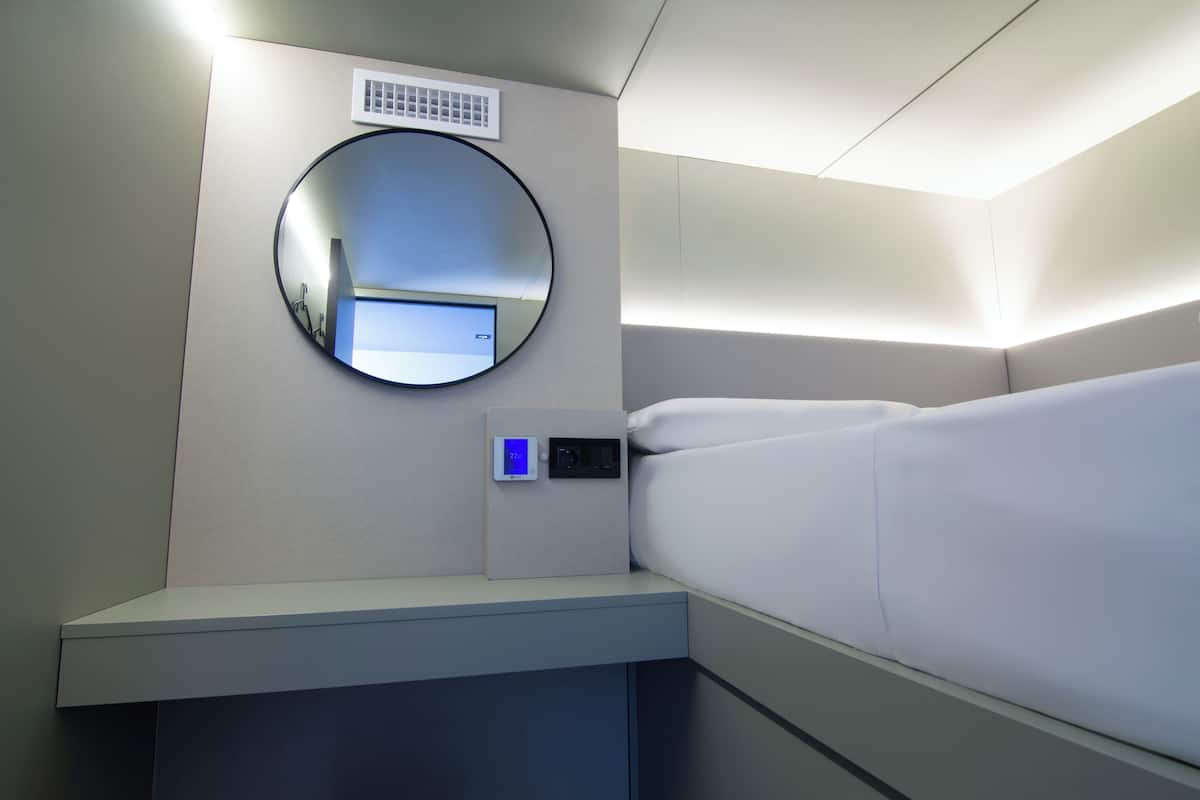 A hotel room with a bed, a bedside table, a digital control panel, a round mirror, and a ventilation unit on the wall.