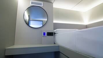A hotel room with a bed, a bedside table, a digital control panel, a round mirror, and a ventilation unit on the wall.