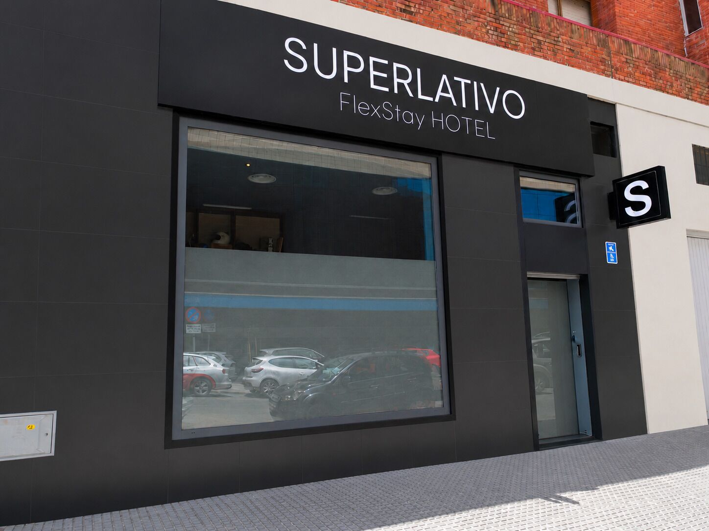 Un bâtiment moderne avec une enseigne indiquant « SUPERLATIVO FlexStay HOTEL » et une grande fenêtre qui reflète les voitures garées.