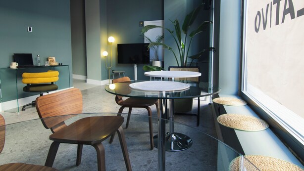 A modern interior with glass tables, wooden chairs, a television, and a large window with 'OVITA' signage. - Hostel Superlativo Vialia I (Málaga)