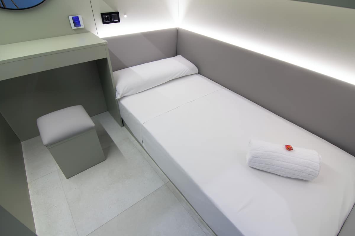 A compact hotel room with a single bed, a small seating area, and a wall-mounted control panel.