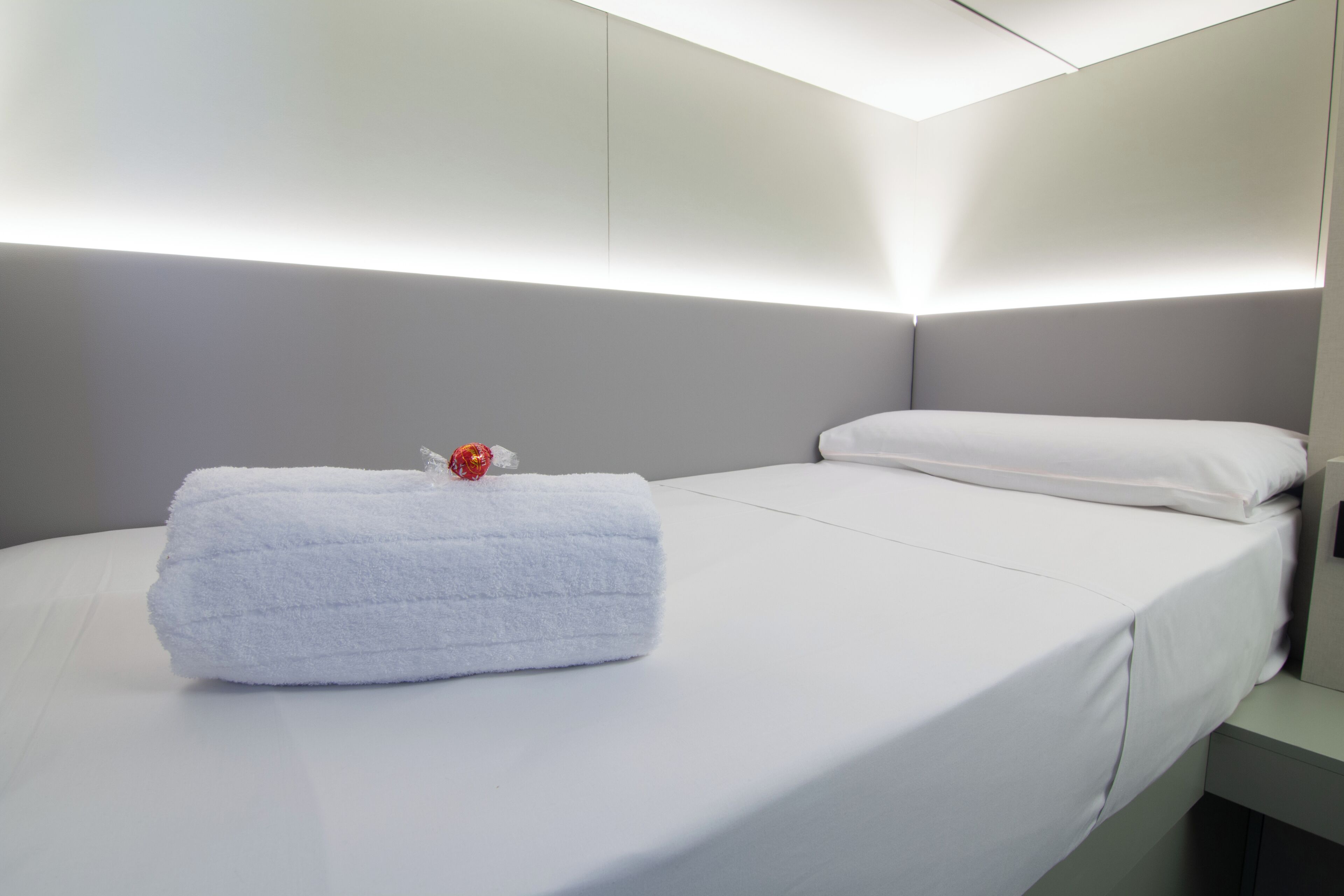 Una cama bien hecha con una toalla blanca y un adorno rojo, un cabecero gris y una pared blanca.