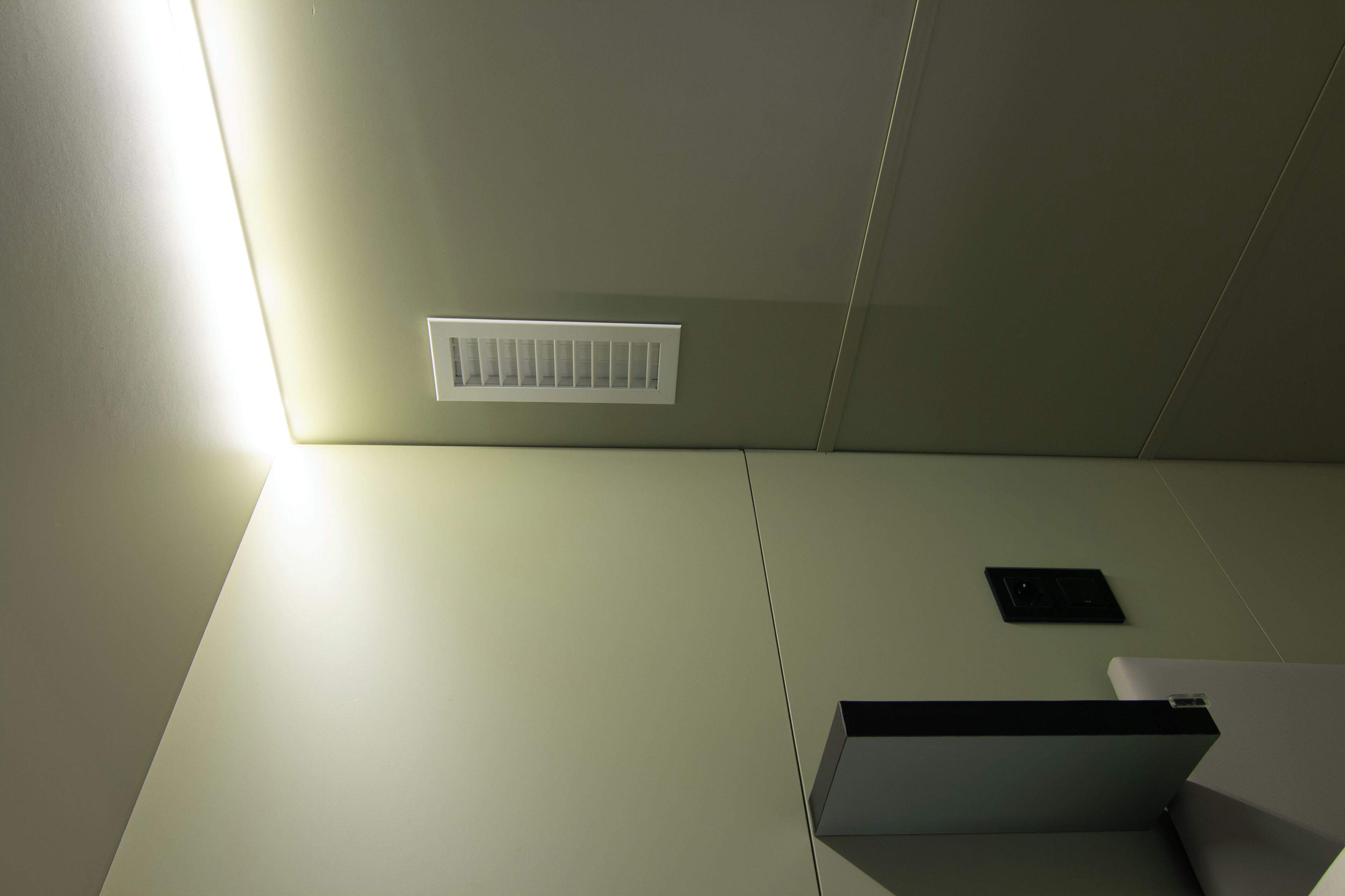 Een kamer met een wit plafond, een ventilatie rooster, een zwart voorwerp aan de muur en een wit object op de vloer.