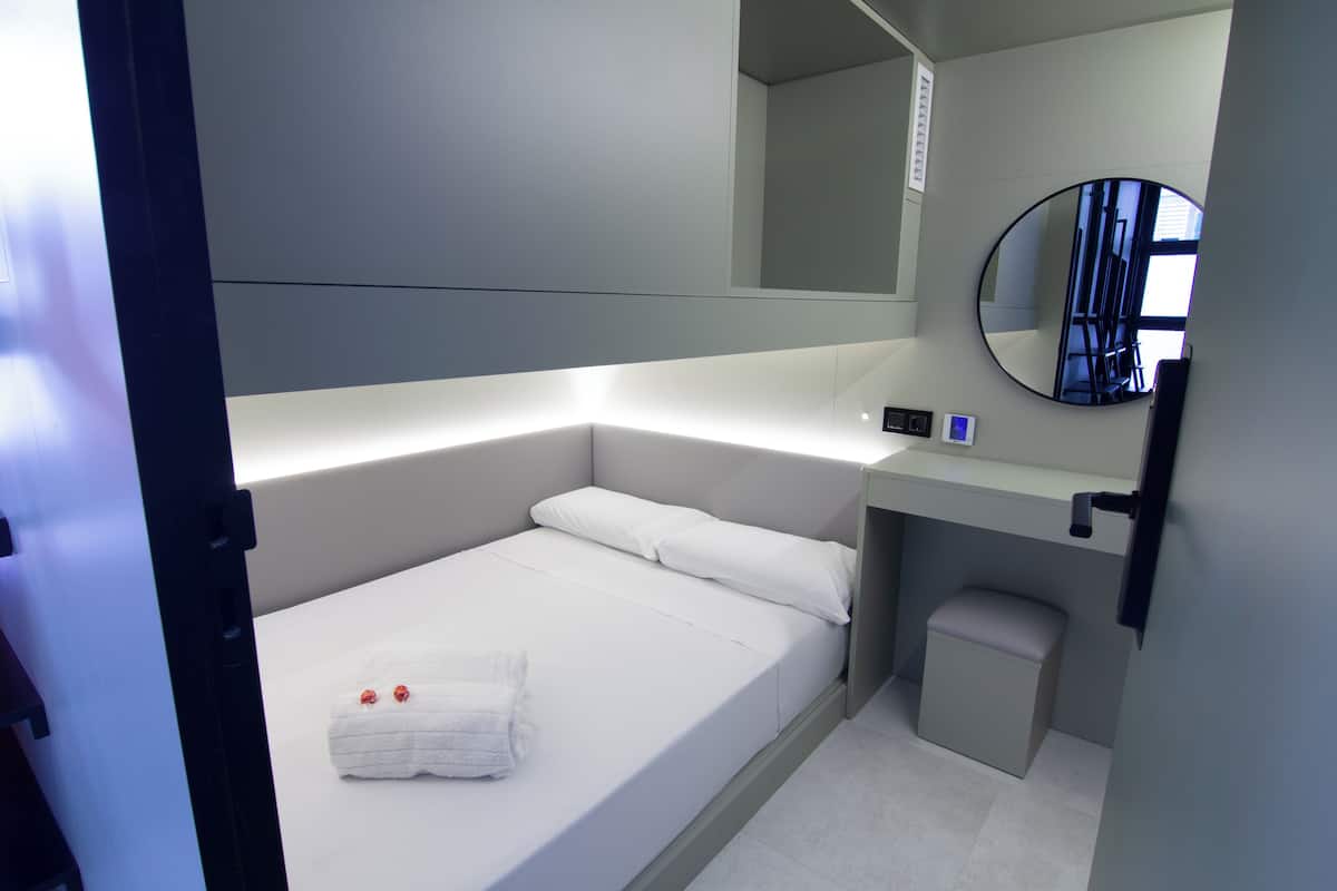 A modern hotel room with a single bed, a bedside table, a mirror, and a small stool.