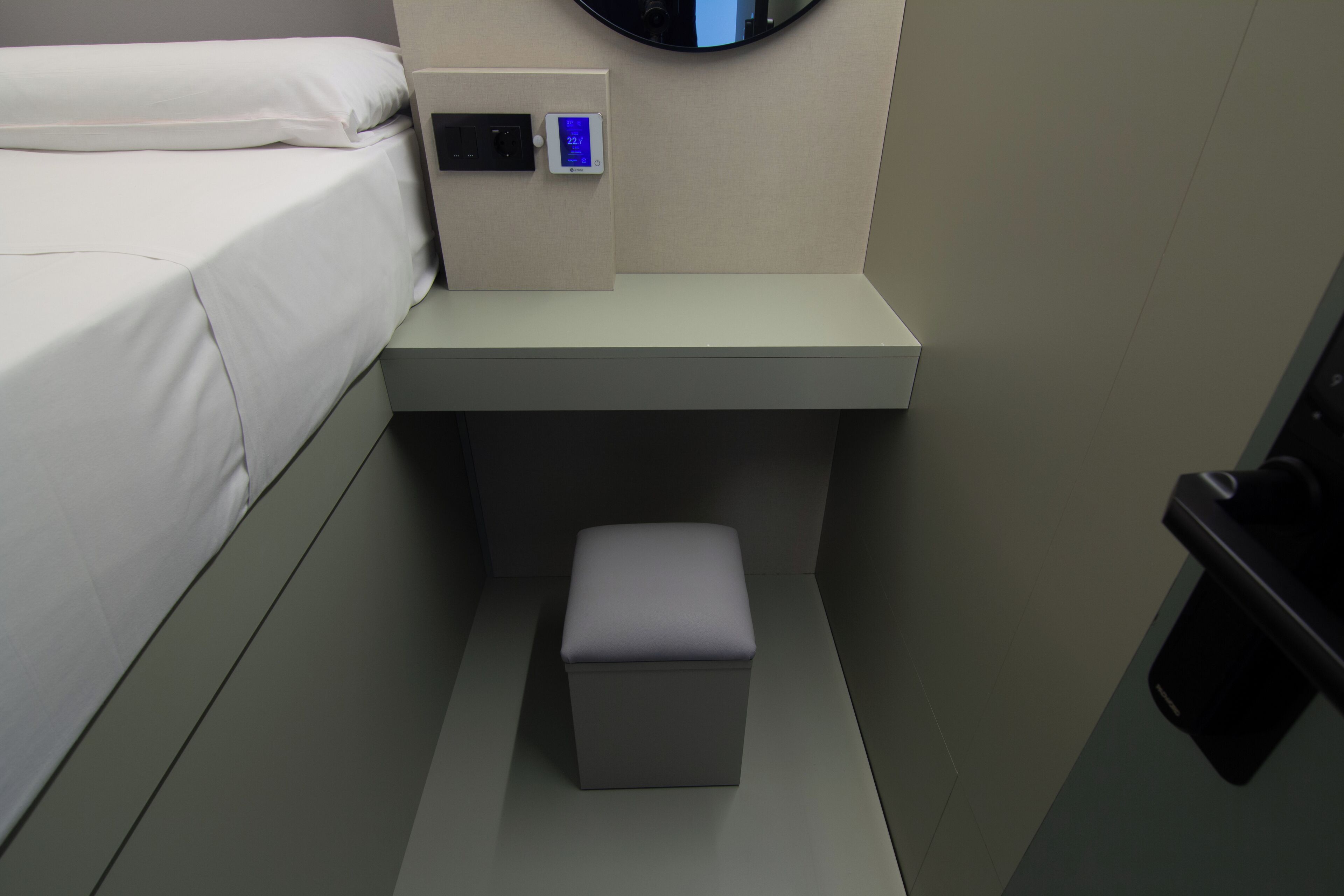 Una habitación de hotel pequeña y moderna con una cama, un panel de control, un taburete pequeño y un espejo en la pared.