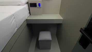 Una habitación de hotel pequeña y moderna con una cama, un panel de control, un taburete pequeño y un espejo en la pared.