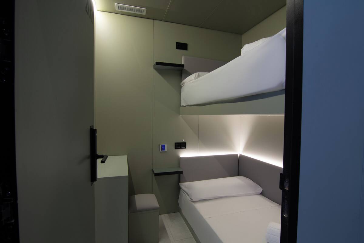 A compact room with bunk beds, a small bedside table, and a wall-mounted light.