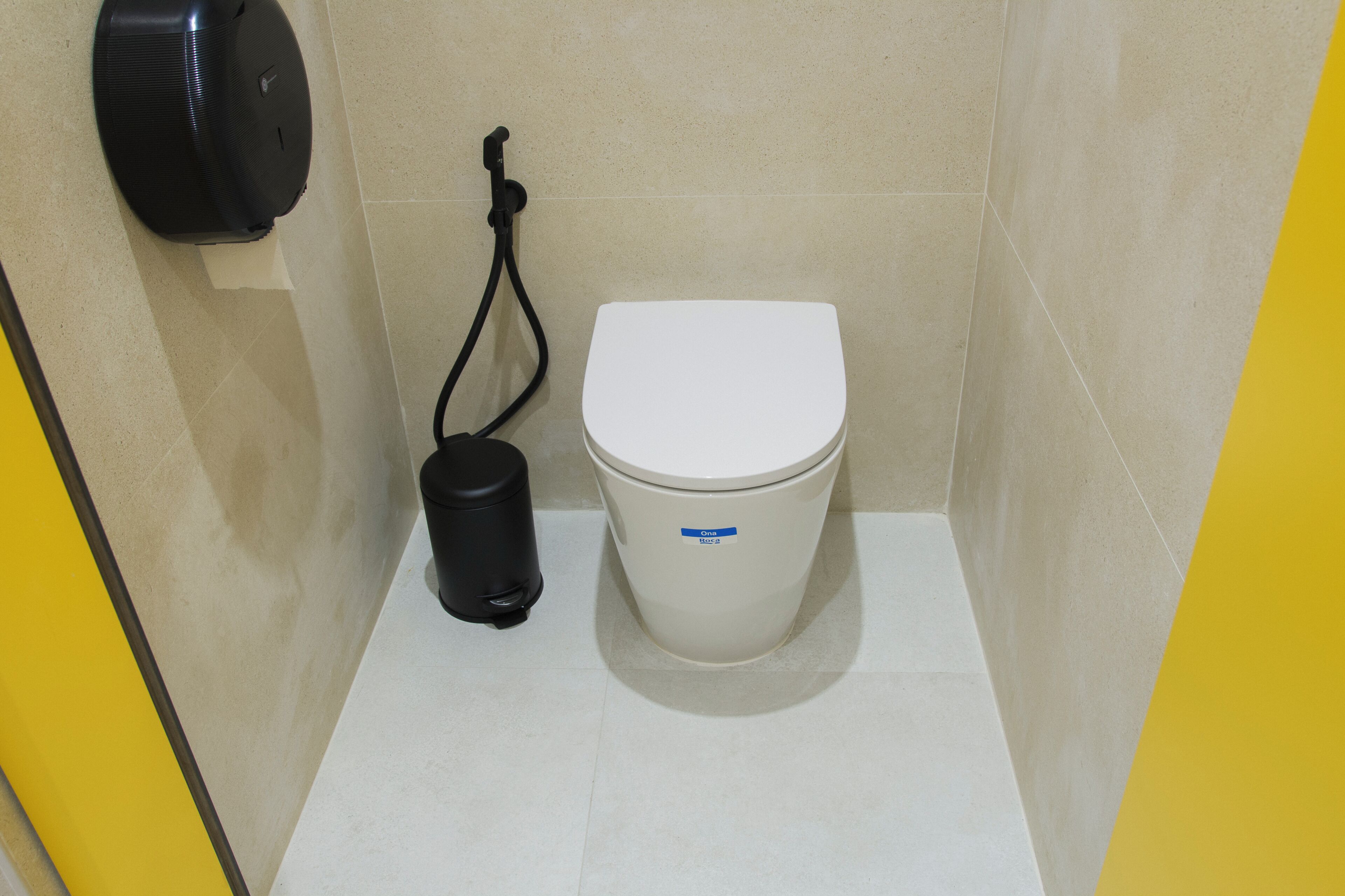 Um banheiro pequeno e limpo, com vaso sanitário branco, lixeira preta e bidê fixado na parede.