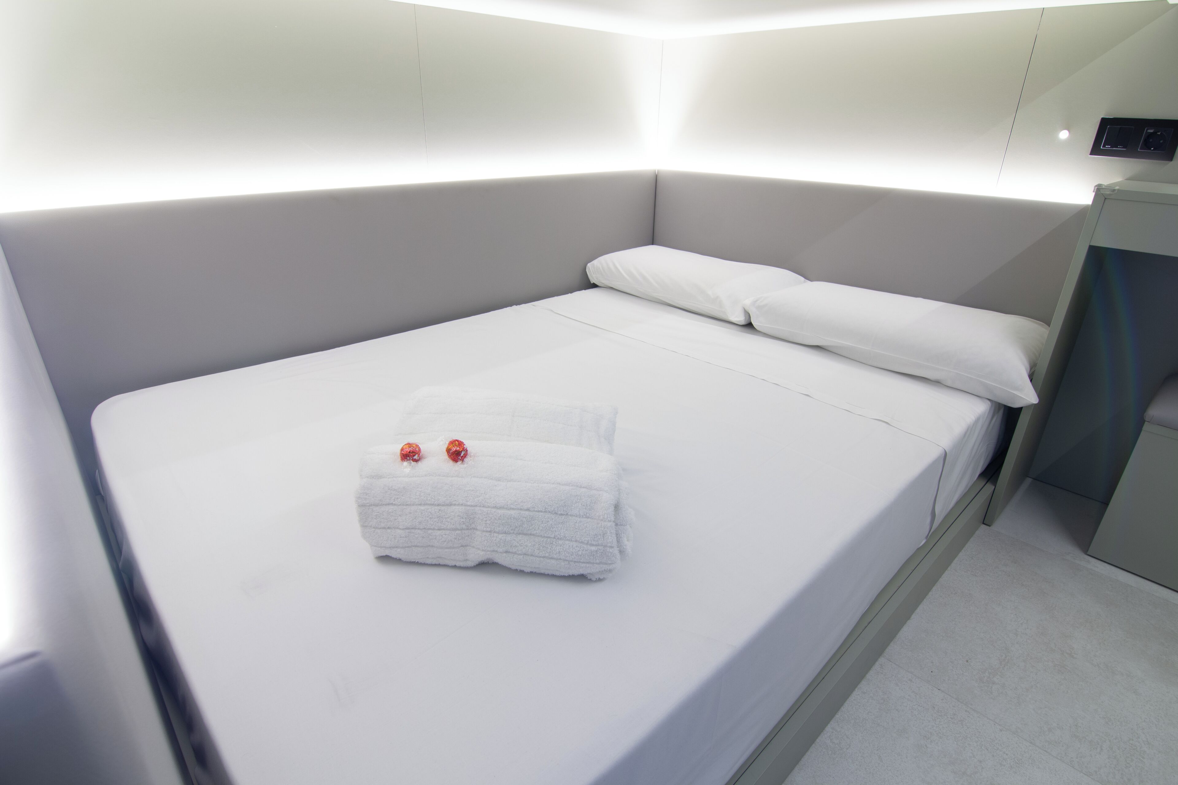 Una cama bien hecha con una toalla blanca doblada y dos adornos rojos, una mesita de noche y un panel de control en la pared.