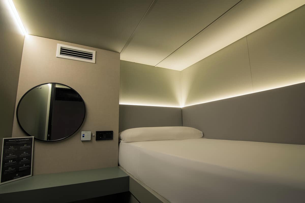 A hotel room with a bed, a round mirror, a wall-mounted air vent, and a small table with a sign.