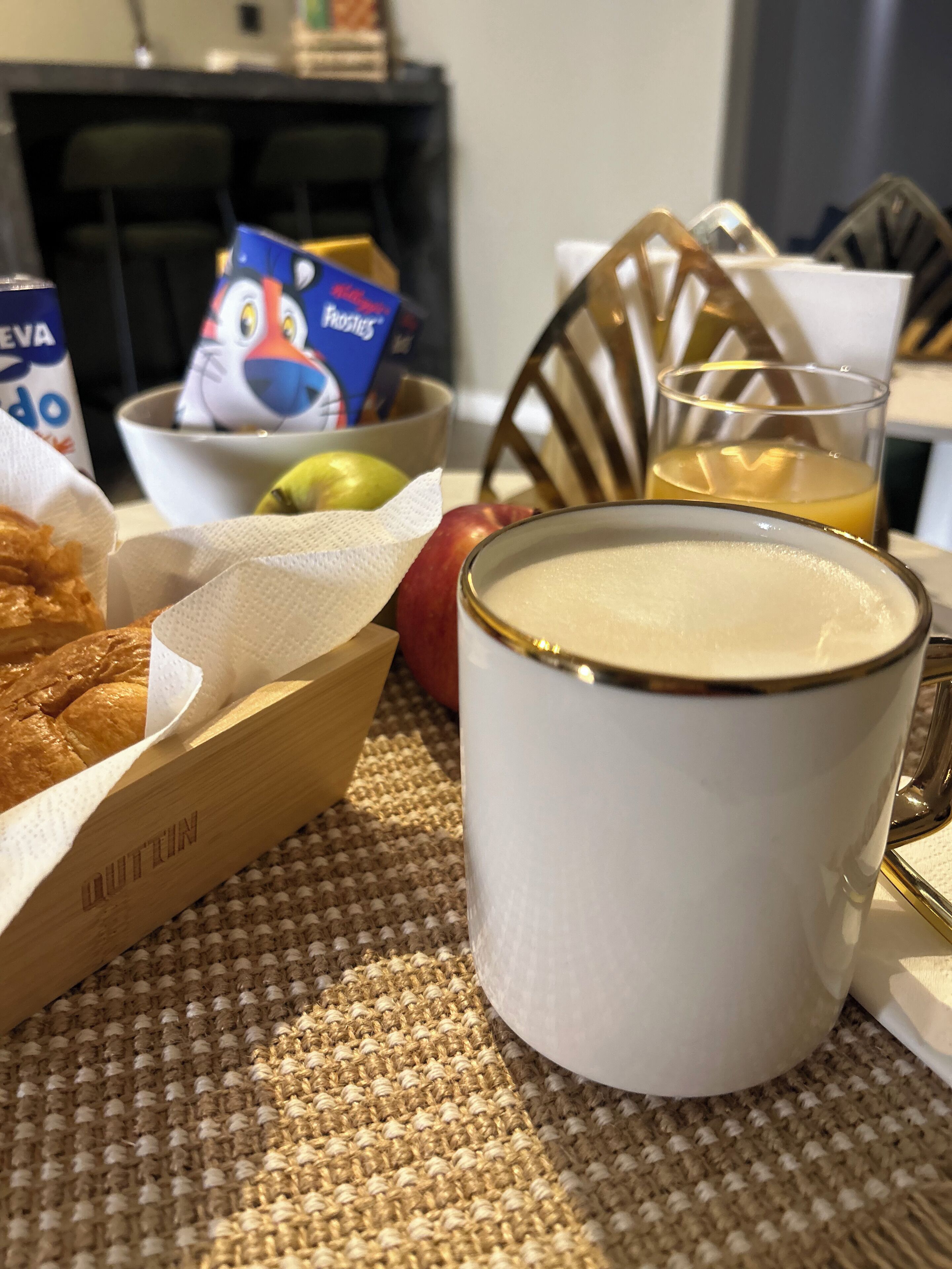 Un desayuno con una taza blanca, un tazón de cereal, una cesta de cruasanes, un vaso de jugo de naranja y una manzana roja sobre un posavasos tejido.