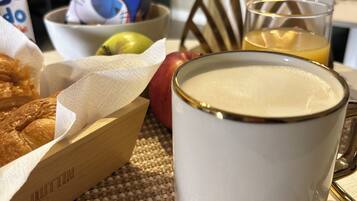 Un desayuno con una taza blanca, un tazón de cereal, una cesta de cruasanes, un vaso de jugo de naranja y una manzana roja sobre un posavasos tejido.