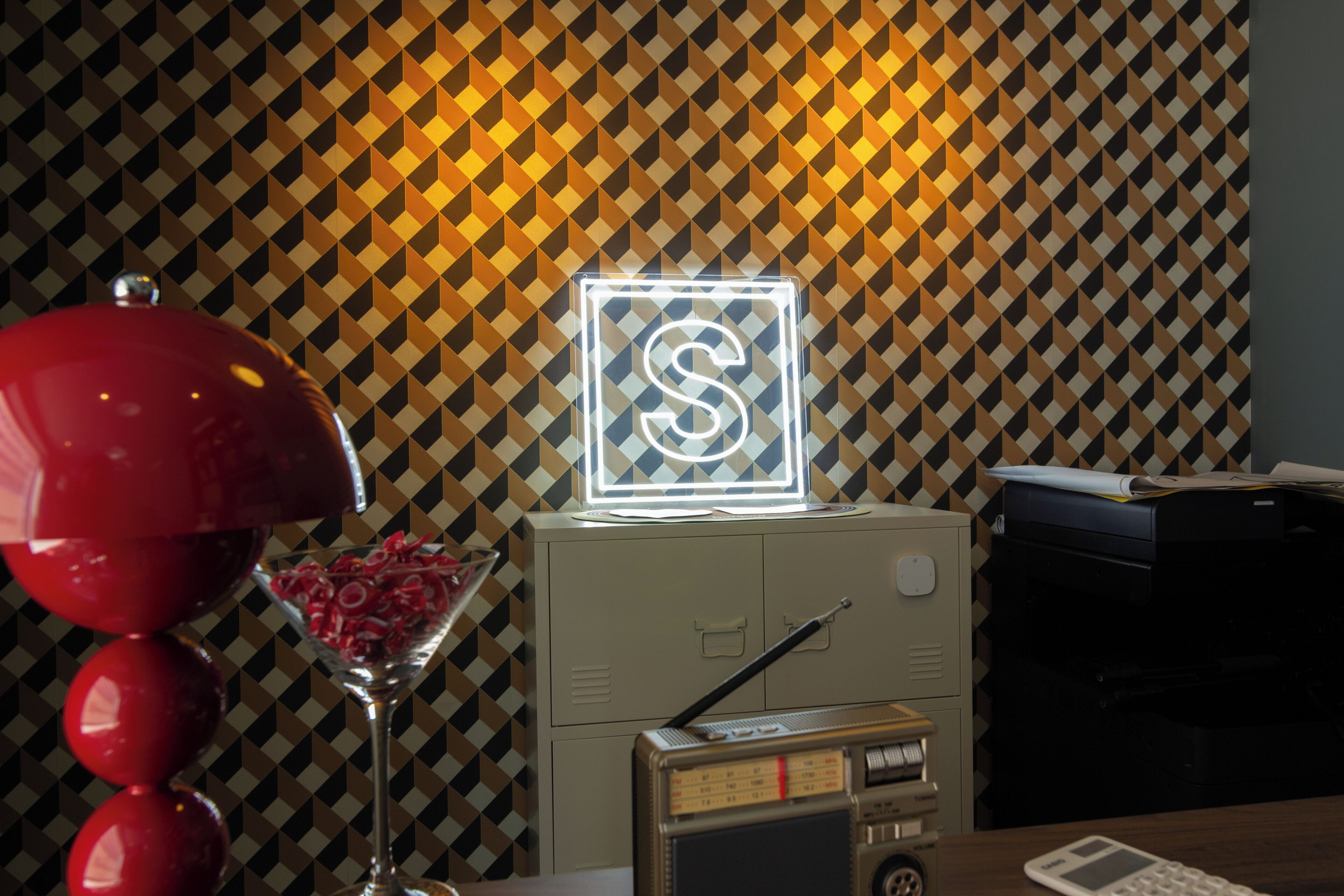A desk with a red lamp, a glass with red items, a neon sign, a radio, and a printer.