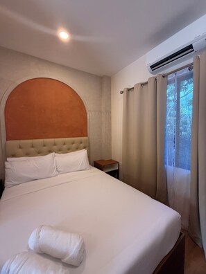 Quarto de hotel com cama, mesa de cabeceira, ar condicionado e janela com cortinas.