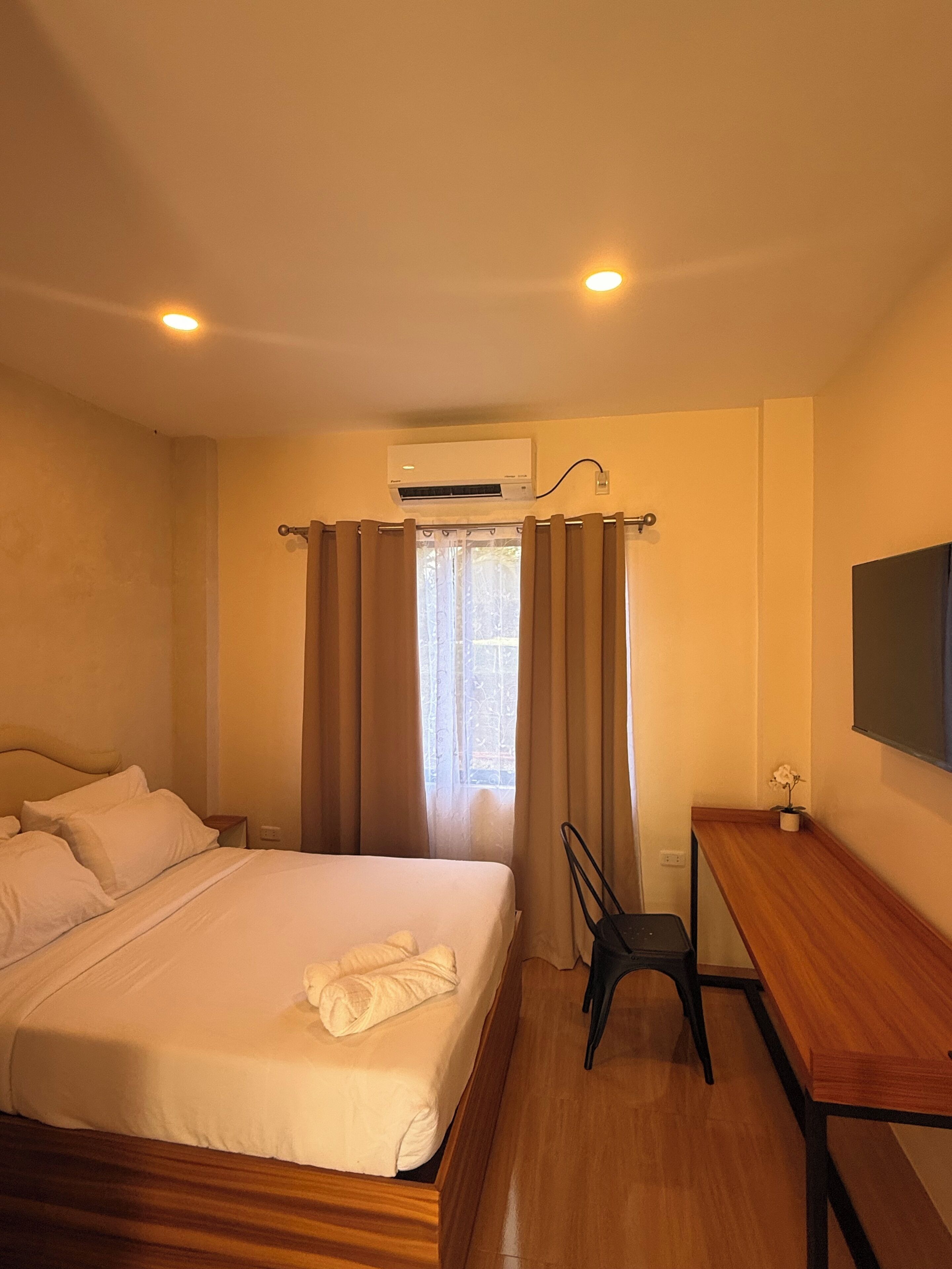 A hotel room with a bed, a desk, a chair, an air conditioning unit, and a window with curtains.