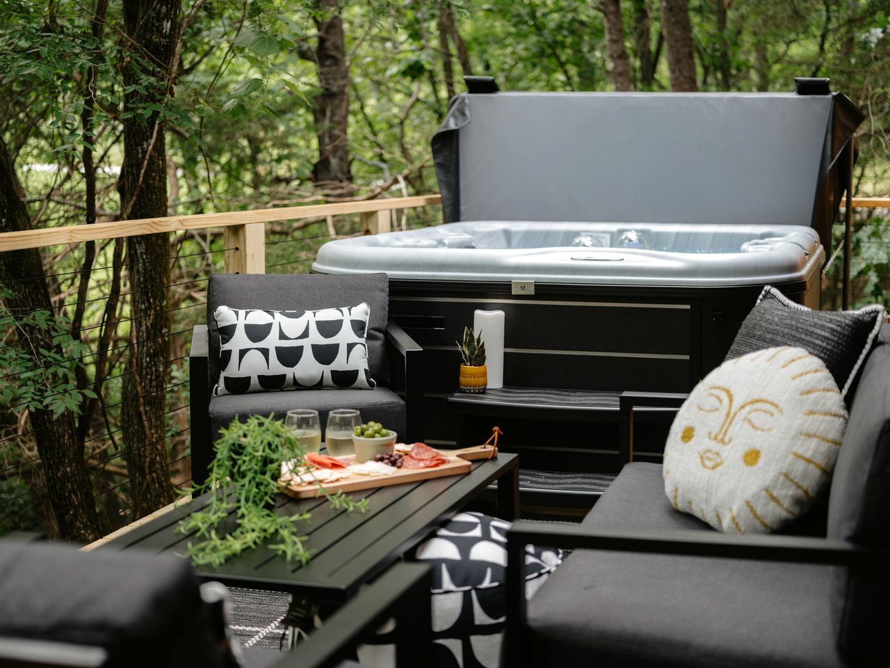 Un espacio exterior acogedor con jacuzzi, una mesa con comida y bebidas, y un sofá con cojines estampados.