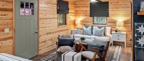 Acogedora habitación de cabaña de madera con cama, dos mesitas de noche, una silla, una pequeña mesa y un ventilador de techo.