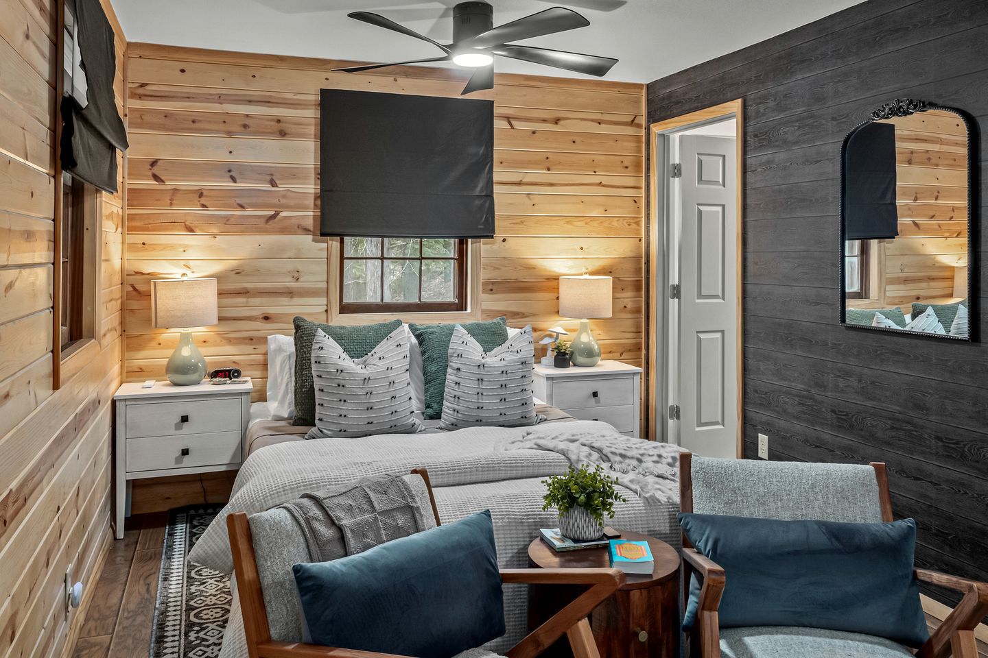 A cozy bedroom with a bed, bedside tables, a desk, chairs, a mirror, and a ceiling fan.