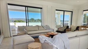 Een moderne woonkamer met een grote bank, een bijzettafeltje en uitzicht op een kustlandschap door grote ramen.