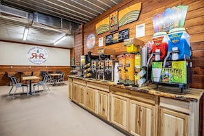 A rustic bar area with multiple beverage dispensers, a logo on the wall, and a seating area with tables and chairs.