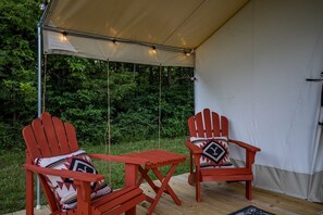 A tent with two red Adirondack chairs and a small table, set on a wooden deck with a view of dense greenery.