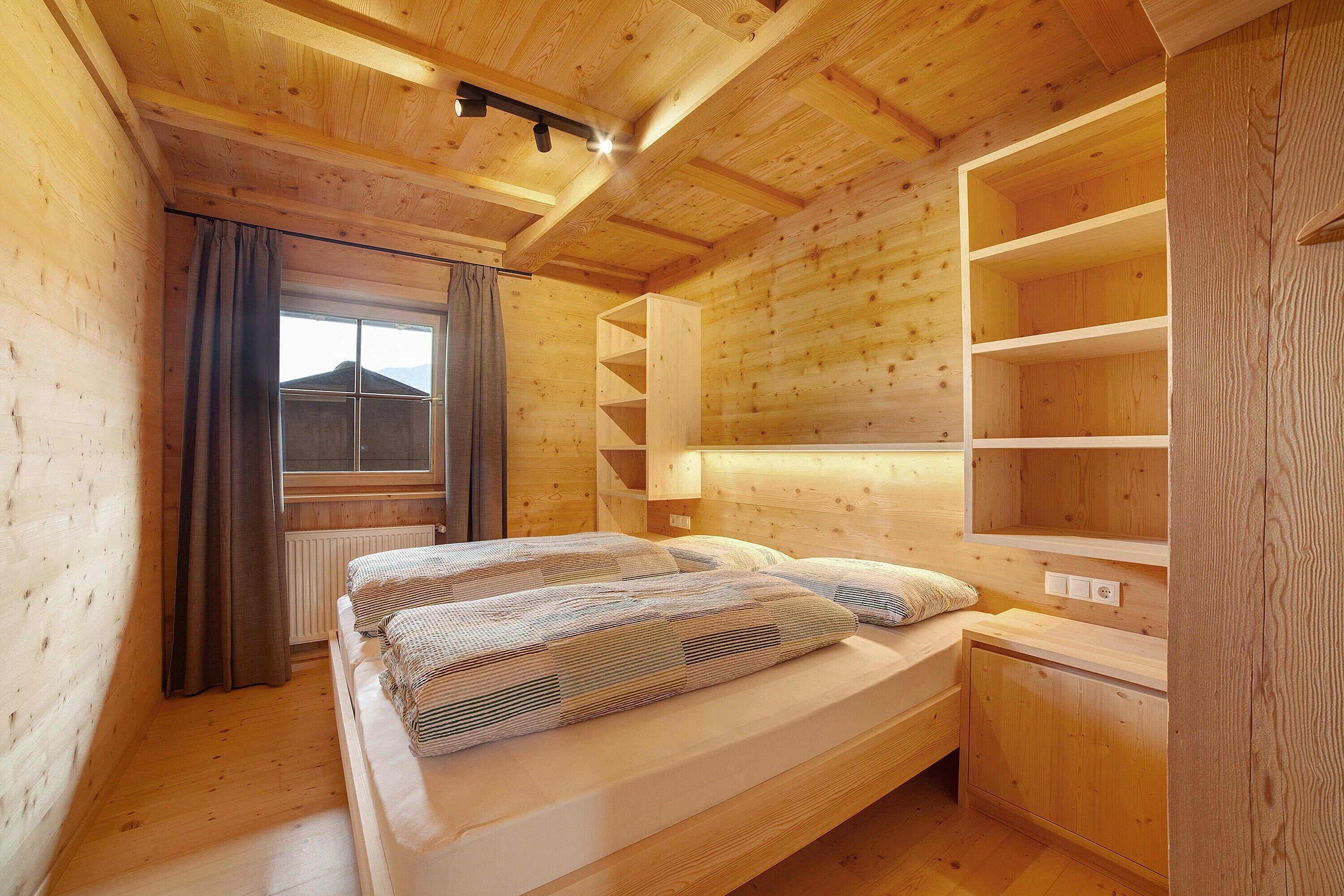 Ein Schlafzimmer einer Holzhütte mit einem Bett, einem Fenster mit Vorhängen und eingebauten Regalen.