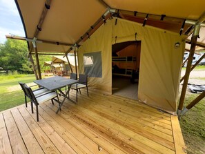 Une tente avec une table et des chaises à l’extérieur, un espace abrité avec un banc, et un véhicule en arrière-plan.