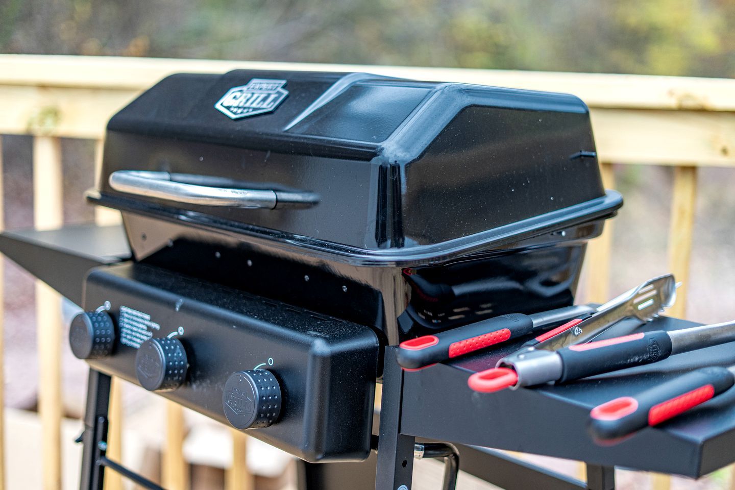 A black grill with a logo, two control knobs, and red-handled tools on a metal stand.