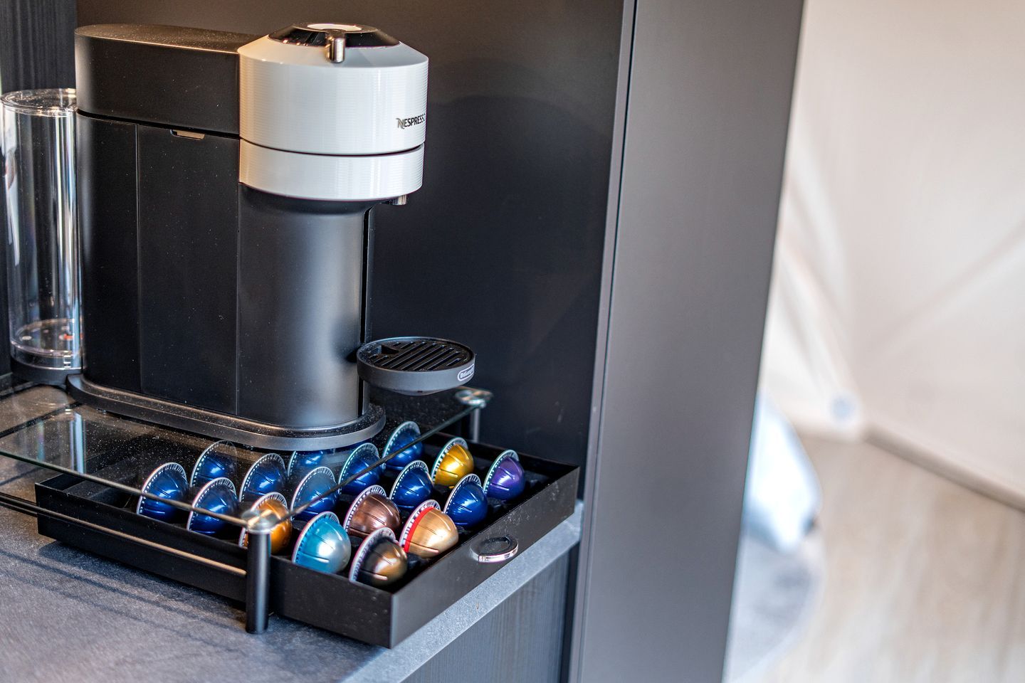 Una cafetera con un surtido de cápsulas de colores, un recipiente de vidrio y una placa metálica.