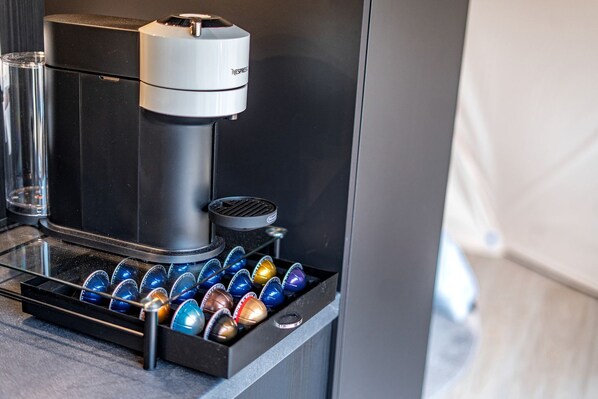 A coffee machine with a tray of colorful capsules, a glass container, and a metal plate.
