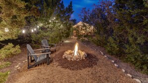 A fire pit with chairs and string lights in a forested area.