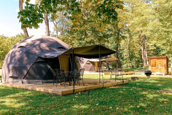 Une tente avec une table et des chaises, un barbecue et une structure en bois, le tout installé dans un espace herbeux bordé d’arbres.