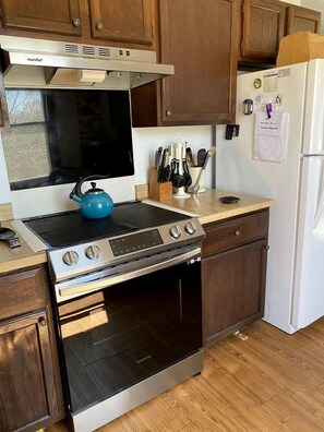 Cocina compacta con estufa, horno y microondas integrados. Un mueble de madera sobre la estufa, un refrigerador a la derecha y una encimera con utensilios.