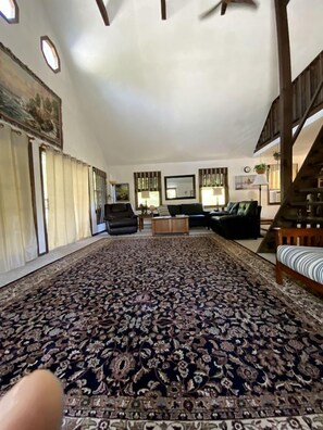 A spacious living room with a large patterned carpet, a staircase, and a view of the outdoors. - Cottage Rental on Chappaquiddick Island with Transferable Ferry Ticket Availability Massachusetts (Edgartown)