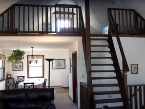 A spacious living area with a dining table, chairs, a black leather sofa, a staircase, and a hanging plant. - Cottage Rental on Chappaquiddick Island with Transferable Ferry Ticket Availability Massachusetts (Edgartown)