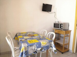 Una habitación pequeña con una mesa cubierta con un mantel estampado, sillas blancas, un microondas en un estante de madera y un televisor montado en la pared.